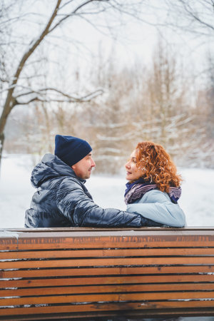 Love romantic love story. Happy couple of middle aged millennials, bright red-haired woman in winter snow park. Romantic date, kissing, hugging. Walking, having fun. Stylish blue clothes, jacket, hat.の写真素材