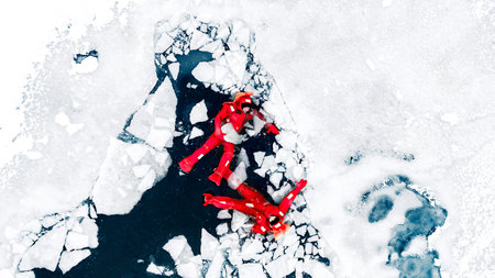 Aerial view of two people ice floating on icy water in bright red survival suits. Arctic frozen landscape. Extreme adventure, ice diving, cold water survival, winter expedition. Fun, outdoor activity.の写真素材