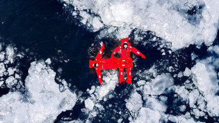 Aerial view of two people ice floating on icy water in bright red survival suits. Arctic frozen landscape. Extreme adventure, ice diving, cold water survival, winter expedition. Fun, outdoor activity.の写真素材