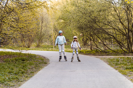 Two girls rollerblading on park path in spring, wearing helmets and knee pads. Children enjoy...