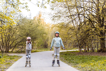 Two girls rollerblading on park path in spring, wearing helmets and knee pads. Children enjoy outdoor sport, combining fun and movement with healthy lifestyle, safety, joy, play and nature outdoorsの写真素材