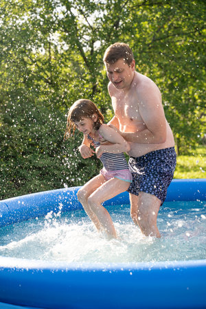 Children playing in inflatable pool on summer day. Two girls laughing, splashing water with father. Family fun, backyard swimming, outdoor activity, bonding, heat relief, happy childhood moment.の写真素材