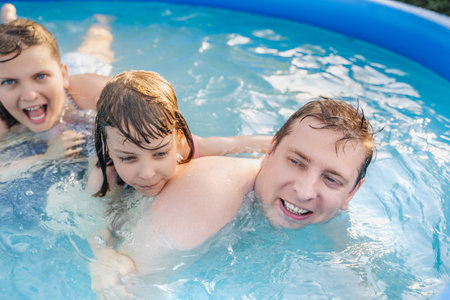 Children playing in inflatable pool on summer day. Two girls laughing, splashing water with father....