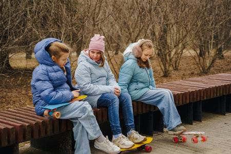 Three girls sit on bench with skateboards in city park. Teens wear jeans, warm jackets, sneakers. Kids rest after outdoor sport, enjoying friendship, balance, street activity and winter freedom.の写真素材