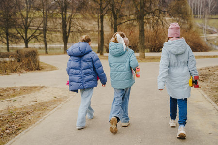 Girls walk with skateboards in city park. Teens wear jackets, jeans, sneakers. Teenagers enjoy street activity and outdoor sport in cold season, building balance, freedom and childhood friendship.の写真素材