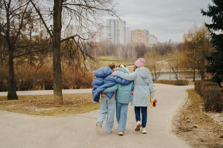 Teen girls walk together, hugging shoulders, each holding a skateboard. Jackets, jeans, sneakers show winter street style. Outdoor friendship, movement, balance, youth bonding, lifestyle and freedom.の写真素材