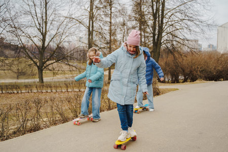 Teen girls ride skateboards on city path in winter park. Jackets and jeans reflect urban youth style. Street sport improves balance, movement, coordination, friendship and outdoor fun for kids.の写真素材