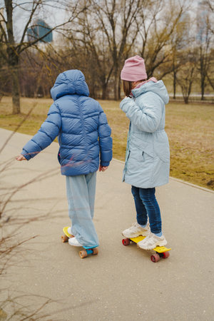 Two girls skate on asphalt path in urban park. Teens in jackets, jeans and sneakers move through cold air. Outdoor street sport builds balance, freedom, energy, motion, fun and childhood friendship.の写真素材