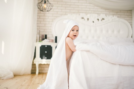 newborn baby five month old baby in the bedroom next to a large white bed on the wooden floor wrapped in a white bamboo towel. Smiles, joy, loveの写真素材
