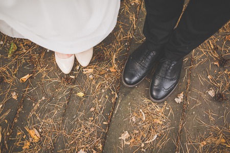 Legs of bride and groom: black shoes, white heeled shoes, details of a wedding gown. On the wooden floor, around the cones and needlesの写真素材