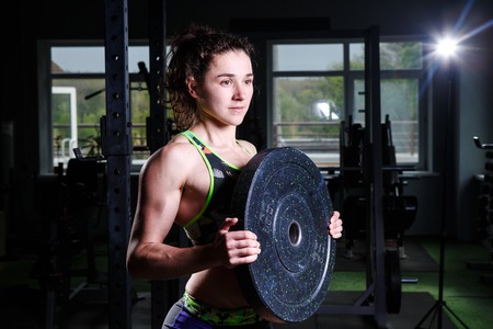Fit power athletic confident young woman crossfit trainer doing exercises with heavy weight barbell plate in gym rising hand. Fitness muscular body, strong hand on dark background Pumping up musclesの写真素材