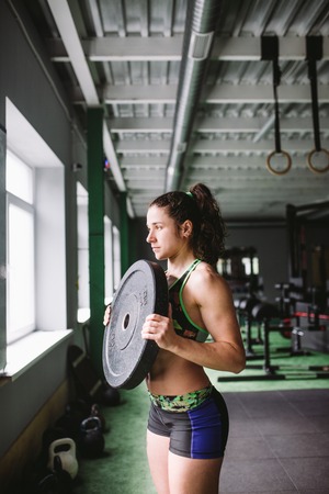 Fit power athletic confident young woman crossfit trainer doing exercises with heavy weight barbell plate in gym rising hand. Fitness muscular body, strong hand on dark background Pumping up musclesの写真素材