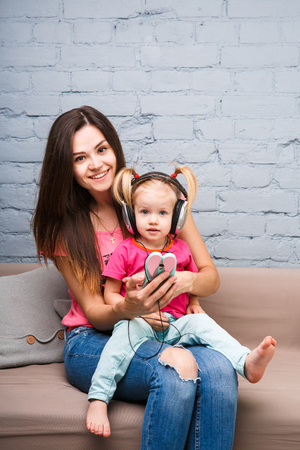 Mom and daughter listen to music in big headphones put on their head, sitting on the sofa in the interior. Holds the phone.の写真素材