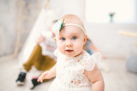 little girl one year with blue eyes blonde in a lush white dress is pleased and plays in a bright room on the background of an Indian tent and parents.の写真素材
