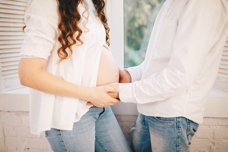 Woman opens her pregnant belly and man puts his hand on it. dressed in white shirts and jeans on the window backgroundの写真素材