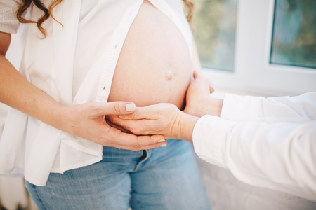 Woman opens her pregnant belly and man puts his hand on it. dressed in white shirts and jeans on the window backgroundの写真素材