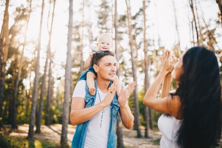 Young beautiful parents playing with their daughter sitting on the shoulders of the pope one year after birth in the coniferous forest in summer.の写真素材