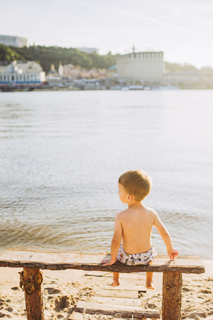 boy child sitting on a wooden bench with his back on the beach near the water and showing his hand forward direction against the backdrop of the city of Kiev.の写真素材