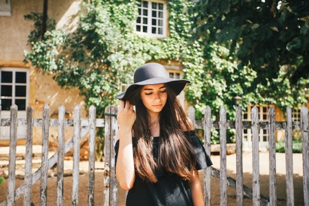 Beautiful sexy girl brunette with brown eyes in a black dress and a black hat with big brims against the backdrop of a wooden fence and an old house in a village, Burgundy in france in summer.の写真素材