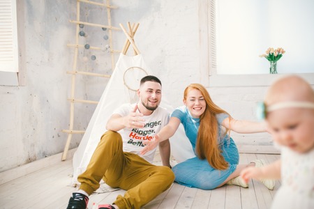 family of three people mom with red hair, dad and daughter begins to walk play in a bright room sitting on a wooden floor against a background of a white Indian tentの写真素材