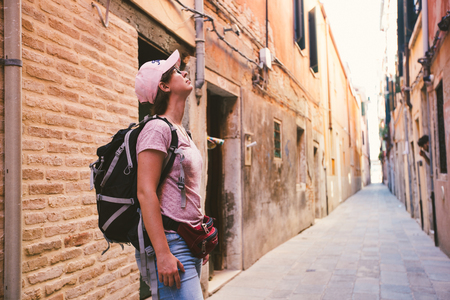 the tourist girl in the caucasian stands on an old deserted street in venice in italy and looks up. Dressed in a pink T-shirt, denim shorts, a cap and a black backpack in the summerの写真素材