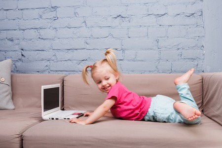Beautiful funny blonde girl a child of two years is lying on the couch indoors and uses a white laptop computer technology with colorful prints pushing the buttons on the keyboard. Dressed in a bright sweatshirt and blue jeansの写真素材