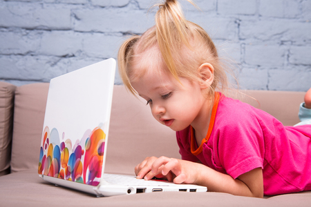 Beautiful funny blonde girl a child of two years is lying on the couch indoors and uses a white laptop computer technology with colorful prints pushing the buttons on the keyboard. Dressed in a bright sweatshirt and blue jeansの写真素材