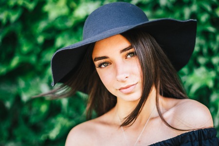 beautiful young sexy girl with swarthy skin and brunette with black hair dressed in a stylish black dress and hat on a background of green foliage.の写真素材