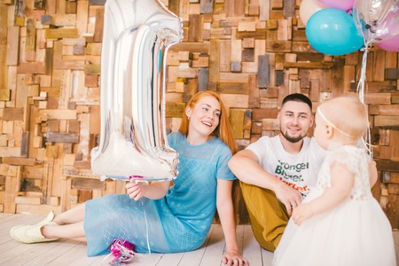 family of three celebrates daughter's birthday one year inside the room sitting on the floor against the background of the wooden derroitive wall. In the hands of holding gel balloons of multi-colored and one in the form of a ballの写真素材