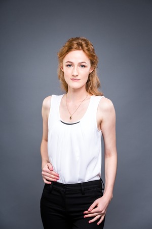 portrait of a young red-haired beautiful girl in the studio on a gray isolated background. A woman is standing with her arms folded and smiling in a white shirt with a short sleeve. Business conceptの写真素材