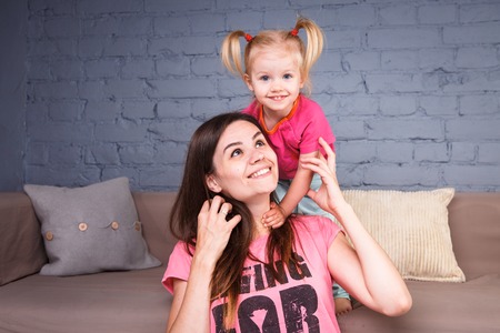 a young slender beautiful mother with a blond daughter on her shoulders play, am happy and indulge in the committees near the sofa on the wooden floor. Family valuesの写真素材
