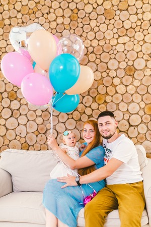 young family man, woman and baby girl sitting on couch indoors with wooden decorative wall and multi-colored balloons with helium in hand. Children's holidayの写真素材