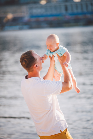 A young father holds one year of his son in his arms with a smile and loves hugs and throws him on the river bank on the beach.の写真素材