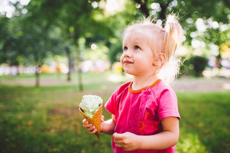 little funny girl blonde eating sweet blue ice cream in a waffle cup on a green summer background in the park. smeared her face and cheeks and laughs. Dressed in bright stylish clothes.の写真素材