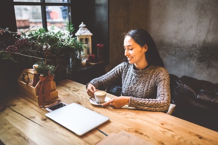 beautiful business girl drinking coffee from white cup in restaurant decorated with Christmas decor.sitting near window at a wooden table.Dressed in warm gray sweater. On the table, phone and glasses.の写真素材