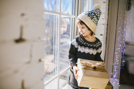 child is sitting by the window on a sunny Christmas day and makes out with gifts in boxes wrapped in paper.dressed in knitted warm woolen clothes and a hat. Inside the house with a stylish interior.の写真素材