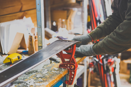 Theme repairs and maintenance of skis. The male worker is repairing work clothes, applying wax on the sliding surface onto skis mounted on ski vise of red color.The concept of service in the workshop.の写真素材