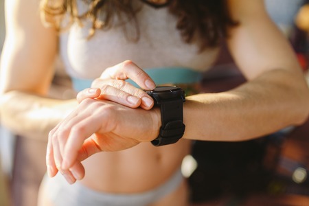 Theme of sport and technology. A strong, beautiful woman with a flat belly near the window on a sunny day presses the buttons, uses a black sports watch on her arm. Close-up.の写真素材
