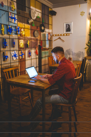 man with beard businessman working behind laptop in coffee shop in evening. interior decorated with Christmas decor, light bulbs are shining, hands on keyboard typing text. The concept small business.の写真素材