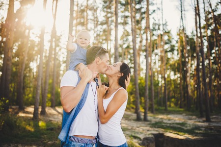 Family mom, dad and daughter sits at the daddy on the shoulders, and parents kiss on the nature in the forest in the summer at sunset.の写真素材