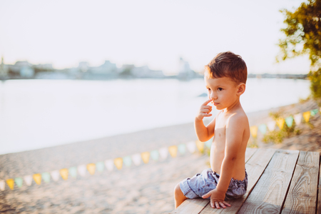 The theme is child and summer beach vacation. A small Caucasian boy sits sideways on a wooden pier and admires the view of the sandy beach and the pond, the river. With bare legs in blue denim shortsの写真素材