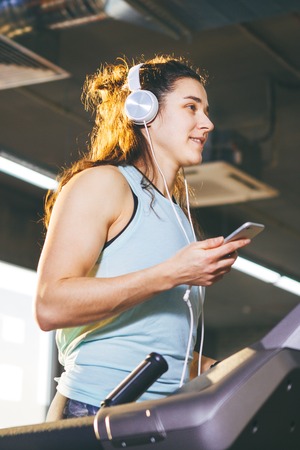 Theme sport and music. A beautiful Caucasian woman running in gym on treadmill. On head big white...