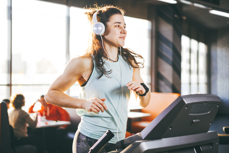 Theme is sport and music. A beautiful inflated woman runs in the gym on a treadmill. On her head are big white headphones, the girl listens to music during a cardio workout for weight loss.の写真素材