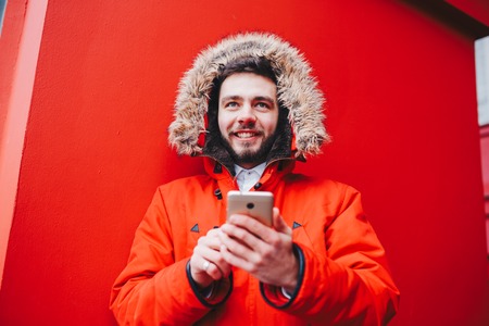 handsome young male student with toothy smile and beard stands on red wall background, facade of educational institution in red winter jacket with hood with fur, Uses finger on screen of mobile phone.の写真素材