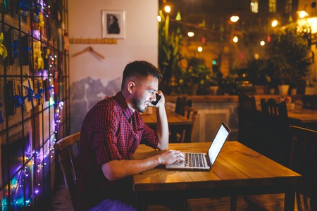 Young handsome Caucasian man with beard and toothy smile in red shirt works behind laptop, hands on keyboard sitting at wooden table. Uses calls on mobile phone. In evening at the coffee shop.の写真素材