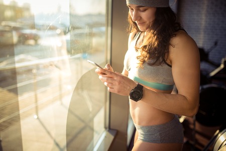A beautiful sexy Caucasian woman stands near a window with sunlight in the gym. Holds in his hand, uses a cell phone. Dressed in gray sportswear and a hat.の写真素材