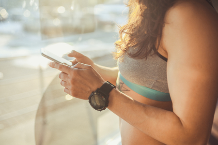 hands Beautiful sexy caucasian woman stands near window with sunlight in the gym. Holds in his hand, uses cell phone and sports watch. Dressed in gray sportswear and a hat. Unrecognizable Personalityの写真素材