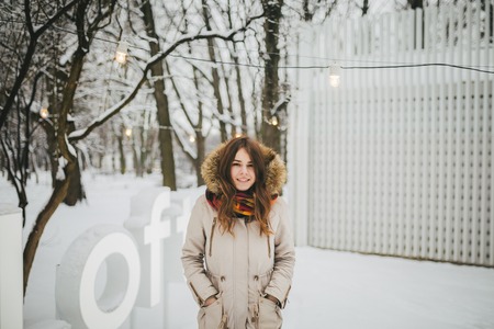 Theme is a weekend holiday in winter. Beautiful young Caucasian woman standing in snow park in jacket with hood and fur in jeans and smiling.の写真素材