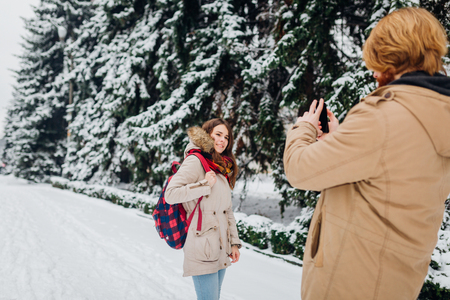 Young couple in love rest in the park winter. A man holds mobile phone in hands and takes a photo of girlfriend against the background of snowcovered trees. Woman in jacket and red backpack.の写真素材
