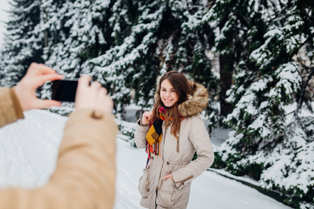 Young couple in love rest in the park winter. A man holds mobile phone in hands and takes a photo of girlfriend against the background of snowcovered trees. Woman in jacket and red backpack.の写真素材